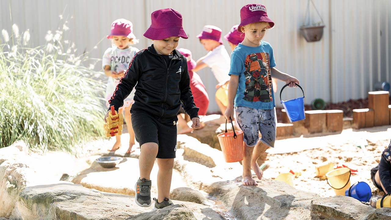 Children walking around a sandpit, carrying buckets and sand toys.